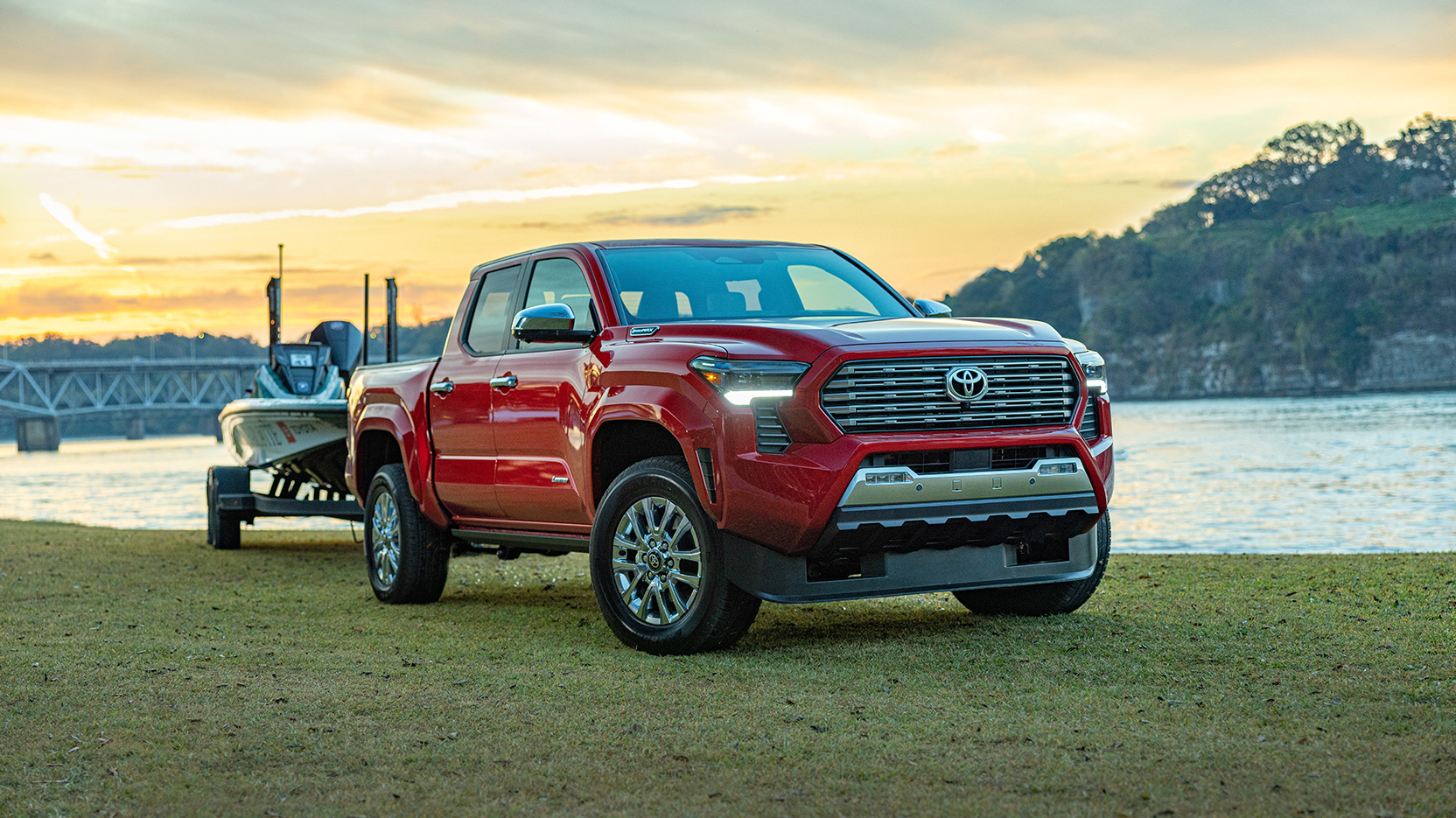 A red Toyota Tacoma towing a bass boat at a lake at sunset